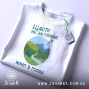 Футболка щастя в горах, біла, складена на поверхні, фон світло-сірий, принт з горами, річкою та деревами в зелених відтінках, текст українською мовою, з етикеткою на шнурку.