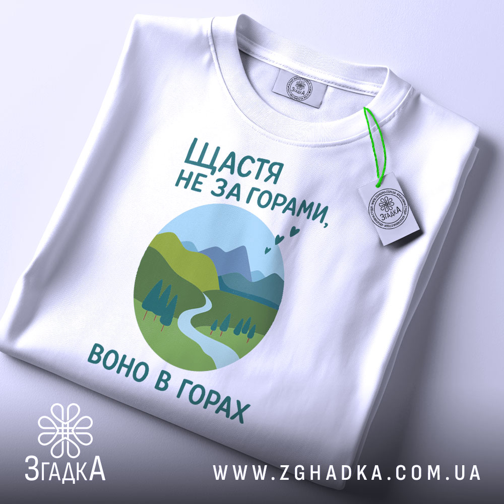 Футболка щастя в горах, біла, складена на поверхні, фон світло-сірий, принт з горами, річкою та деревами в зелених відтінках, текст українською мовою, з етикеткою на шнурку.