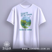 Футболка щастя в горах, біла, підвішена на вішалці, фон світло-сірий, принт з горами, річкою та деревами в зелених відтінках, текст українською мовою.