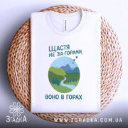 Футболка щастя в горах, біла, складена на плетеній кошику, фон світло-сірий, принт з горами, річкою та деревами в зелених відтінках, текст українською мовою.