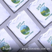 Футболка щастя в горах, біла, зображена на манекені, фон світло-сірий, принт з горами, річкою та деревами в зелених відтінках, текст українською мовою.