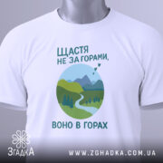Футболка щастя в горах, біла, складена на поверхні, фон світло-сірий, принт з горами, річкою та деревами в зелених відтінках, текст українською мовою, з етикеткою на шнурку.