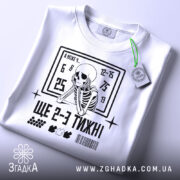 Футболка ще 2-3 тижні, біла, складена на поверхні, фон світло-сірий, принт з чорним скелетом, що сидить, та текстом, на ярлику зелена стрічка.