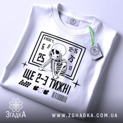 Футболка ще 2-3 тижні, біла, складена на поверхні, фон світло-сірий, принт з чорним скелетом, що сидить, та текстом, на ярлику зелена стрічка.