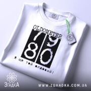 Футболка ще той олдовий. Біла футболка, складена на столі, з видимим етикеткою. Принт чорного кольору з написом 'OLDOMETER 79 80' та фразою 'Я ще той олдовий!' розташований по центру.