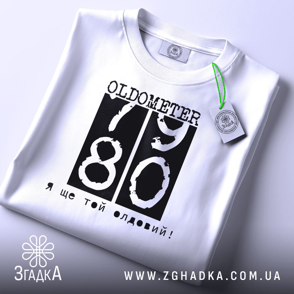 Футболка ще той олдовий. Біла футболка, складена на столі, з видимим етикеткою. Принт чорного кольору з написом 'OLDOMETER 79 80' та фразою 'Я ще той олдовий!' розташований по центру.