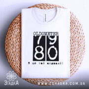 Футболка ще той олдовий. Біла футболка, складена на плетеному кошику, з видимим принтом чорного кольору з написом 'OLDOMETER 79 80' та фразою 'Я ще той олдовий!' внизу.