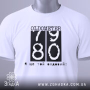 Футболка ще той олдовий. Біла футболка, складена на столі, з видимим етикеткою. Принт чорного кольору з написом 'OLDOMETER 79 80' та фразою 'Я ще той олдовий!' розташований по центру.