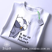 Футболка що це ви робите. Біла футболка, складена на поверхні, з принтом птаха, що виглядає з-за стіни, текстом 'А що це Ви тут Робите?'. Вид з верхнього ракурсу, фон світлий.