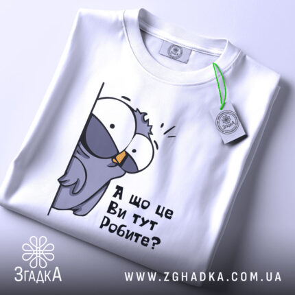 Футболка що це ви робите. Біла футболка, складена на поверхні, з принтом птаха, що виглядає з-за стіни, текстом 'А що це Ви тут Робите?'. Вид з верхнього ракурсу, фон світлий.