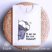 Футболка що це ви робите. Біла футболка, складена на плетеній кошику, з принтом птаха, що виглядає з-за стіни, текстом 'А що це Ви тут Робите?'. Вид з верхнього ракурсу, фон світлий.