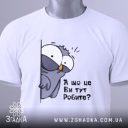 Футболка що це ви робите. Біла футболка, складена на поверхні, з принтом птаха, що виглядає з-за стіни, текстом 'А що це Ви тут Робите?'. Вид з верхнього ракурсу, фон світлий.