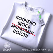 Футболка білого кольору, зображена зверху, на ній видно принт чорного кольору з текстом "ПОТРІБНО ЩОСЬ ЗМІНЮВАТИ ПОЇСТИ", на комірці є етикетка, фон світло-сірий.