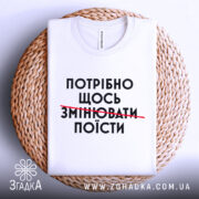 Футболка білого кольору, зображена зверху на плетеній підставці, фон світло-сірий, принт чорного кольору з текстом "ПОТРІБНО ЩОСЬ ЗМІНЮВАТИ ПОЇСТИ".