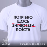 Футболка білого кольору, зображена зверху, на ній видно принт чорного кольору з текстом "ПОТРІБНО ЩОСЬ ЗМІНЮВАТИ ПОЇСТИ", на комірці є етикетка, фон світло-сірий.