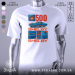 Футболка Shelby GT білого кольору, зображена на манекені, ракурс спереду, фон світло-сірий, принт містить зображення автомобіля Shelby GT500 та текст "500 SHELBY" в червоному та синьому кольорах.