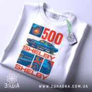 Футболка Shelby GT білого кольору, зображена на столі, ракурс зверху, фон світло-сірий, принт містить зображення автомобіля Shelby GT500 та текст "500 SHELBY" в червоному та синьому кольорах, на футболці є етикетка.