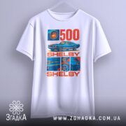 Футболка Shelby GT білого кольору, зображена на вішалці, ракурс спереду, фон світло-сірий, принт містить зображення автомобіля Shelby GT500 та текст "500 SHELBY" в червоному та синьому кольорах.