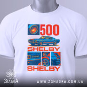 Футболка Shelby GT білого кольору, зображена на столі, ракурс зверху, фон світло-сірий, принт містить зображення автомобіля Shelby GT500 та текст "500 SHELBY" в червоному та синьому кольорах, на футболці є етикетка.