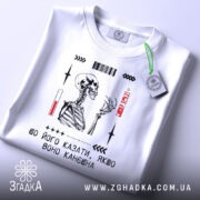 Футболка шо його казати, біла, зображена з близького ракурсу, лежить на поверхні, фон світло-сірий, принт з малюнком скелета, текстом та етикеткою зеленого кольору.