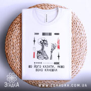 Футболка шо його казати, біла, зображена зверху, лежить на плетеній кошику, фон світло-сірий, принт з малюнком скелета та текстом у чорному і червоному кольорах.