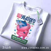 Футболка шубатого нового року, білого кольору, зображена зверху, фон світлий, принт з рибою в рожевій шубі, на футболці є етикетка, яскраві кольори, деталі принта включають сердечка та дорогоцінні камені.