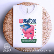 Футболка шубатого нового року, білого кольору, зображена зверху на плетеній основі, фон світлий, принт з рибою в рожевій шубі, яскраві кольори, деталі принта включають сердечка та дорогоцінні камені.