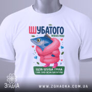 Футболка шубатого нового року, білого кольору, зображена зверху, фон світлий, принт з рибою в рожевій шубі, на футболці є етикетка, яскраві кольори, деталі принта включають сердечка та дорогоцінні камені.