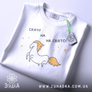 Футболка скачу на свято, біла, складена на столі, фон світло-сірий, принт з єдинорогом та текстом 'скачу на свято!', з зеленим ярликом.