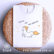 Футболка скачу на свято, біла, складена на плетеній кошику, фон світло-сірий, принт з єдинорогом та текстом 'скачу на свято!'.