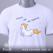 Футболка скачу на свято, біла, складена на столі, фон світло-сірий, принт з єдинорогом та текстом 'скачу на свято!', з зеленим ярликом.