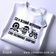 Футболка скажений агроном, біла, складена на поверхні, фон світлий, принт чорного кольору з текстом і іконками, що зображують агронома, пиво та трактор, з етикеткою на комірці.