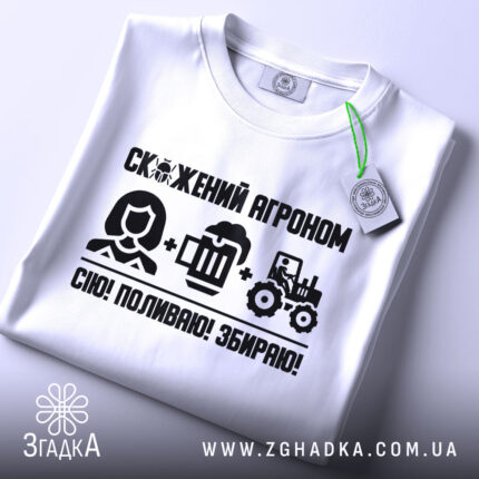 Футболка скажений агроном, біла, складена на поверхні, фон світлий, принт чорного кольору з текстом і іконками, що зображують агронома, пиво та трактор, з етикеткою на комірці.