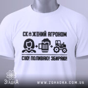 Футболка скажений агроном, біла, складена на поверхні, фон світлий, принт чорного кольору з текстом і іконками, що зображують агронома, пиво та трактор, з етикеткою на комірці.