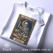 Футболка скелет читає, біла, складена на поверхні, фон світло-сірий, принт з скелетом, що читає книгу, прикрашений квітами та місяцем, з етикеткою на комірці.