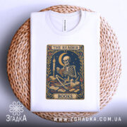 Футболка скелет читає, біла, складена на плетеній кошику, фон білий, принт з скелетом, що читає книгу під місяцем, оточений книгами та квітами.