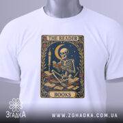 Футболка скелет читає, біла, складена на поверхні, фон світло-сірий, принт з скелетом, що читає книгу, прикрашений квітами та місяцем, з етикеткою на комірці.