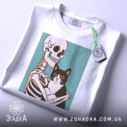 Футболка скелет кіт білого кольору, зображена зверху, на ній видно етикетку, фон світло-сірий, принт зображає скелет, що обіймає чорного кота з великими очима.