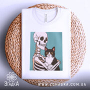 Футболка скелет кіт білого кольору, зображена зверху на плетеному кошику, фон білий, принт зображає скелет, що тримає чорного кота з великими очима.