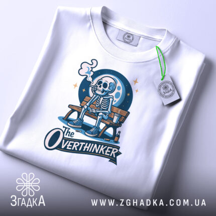 Футболка скелет овердумер, біла, з верхнього ракурсу, на плоскій поверхні, фон світло-сірий, принт зображає скелета на лавці з димом, з написом 'The Overthinker', з етикеткою на комірці.