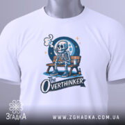 Футболка скелет овердумер, біла, з верхнього ракурсу, на плоскій поверхні, фон світло-сірий, принт зображає скелета на лавці з димом, з написом 'The Overthinker', з етикеткою на комірці.