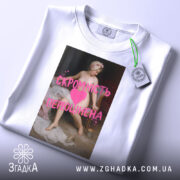 Футболка скромність переоцінена, біла, лежить на столі, фон світлий, принт з класичним живописом та текстом рожевого кольору, на комірці є етикетка.