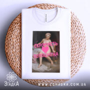 Футболка скромність переоцінена, біла, лежить на плетеній кошику, фон світлий, принт з класичним живописом та текстом рожевого кольору.