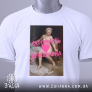 Футболка скромність переоцінена, біла, лежить на столі, фон світлий, принт з класичним живописом та текстом рожевого кольору, на комірці є етикетка.