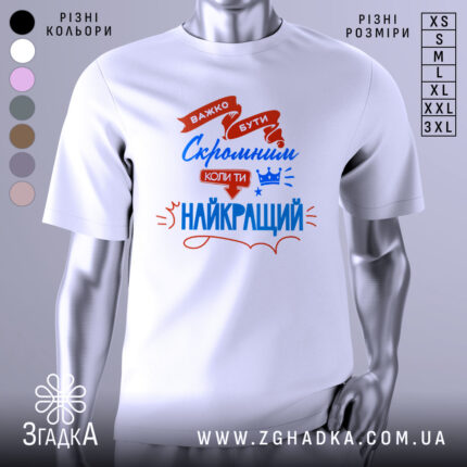 Футболка скромний найкращий. Біла футболка з коротким рукавом, зображена з фронтального ракурсу на манекені. На фоні світло-сірий градієнт. Принт містить червоні та сині елементи з текстом, що говорить про важливість скромності.