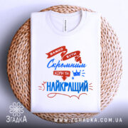 Футболка скромний найкращий. Біла футболка, складена на плоскій поверхні з плетеного матеріалу. На фоні світло-сірий градієнт. Принт з червоними та синіми елементами, текст підкреслює важливість скромності.