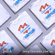 Футболка скромний найкращий. Біла футболка з коротким рукавом, зображена з фронтального ракурсу на манекені. На фоні світло-сірий градієнт. Принт містить червоні та сині елементи з текстом, що говорить про важливість скромності.