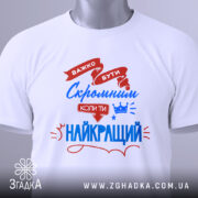 Футболка скромний найкращий. Біла футболка, складена на плоскій поверхні, з видимим етикеткою. На фоні світло-сірий градієнт. Принт з червоними та синіми елементами, текст підкреслює важливість скромності.