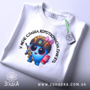 Футболка слабка пам`ять, біла, складена на поверхні з верхнього ракурсу, фон світлий, принт з персонажем у військовій касці, з автоматом та банкою напою, на комірці є етикетка.