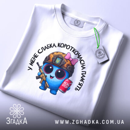Футболка слабка пам`ять, біла, складена на поверхні з верхнього ракурсу, фон світлий, принт з персонажем у військовій касці, з автоматом та банкою напою, на комірці є етикетка.